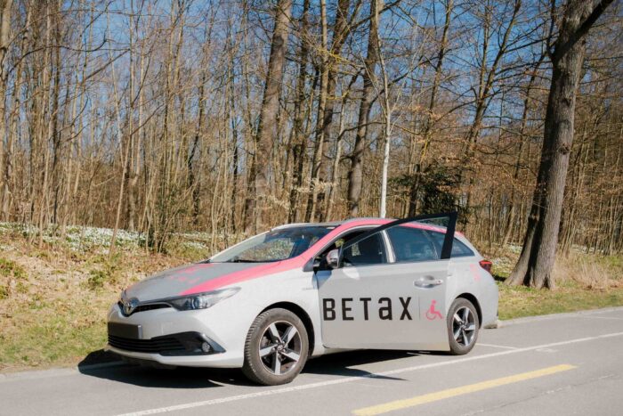 Ein Betax-Personenwagen steht auf einem sonnigen Parkplatz am Waldrand. Die Fahrertür ist geöffnet. Die Bäume haben noch keine Blätter aber erste Frühblüher sind am Waldboden zu sehen.