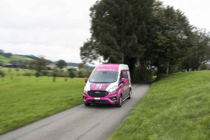 Pink-Weisses Fahrzeug auf Landstrasse. Grüne Wiesen und Bäume im Hintergrund